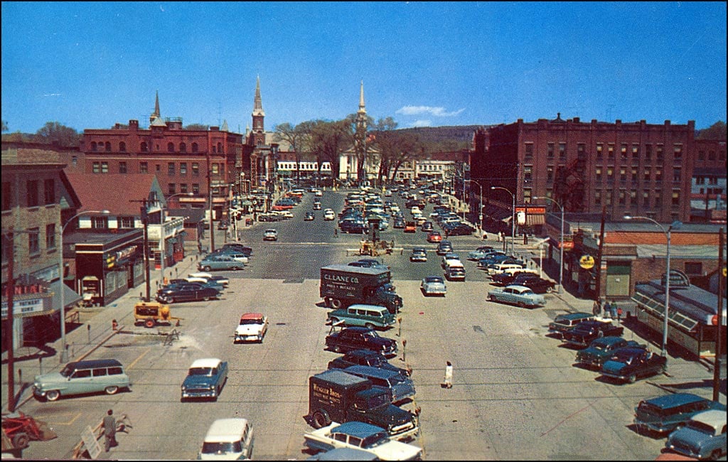 A busy intersection in a small city with vintage cars, brick buildings, traffic lights, and church steeples in the background under a bright blue sky. People are crossing the street and trees are visible in the distance.