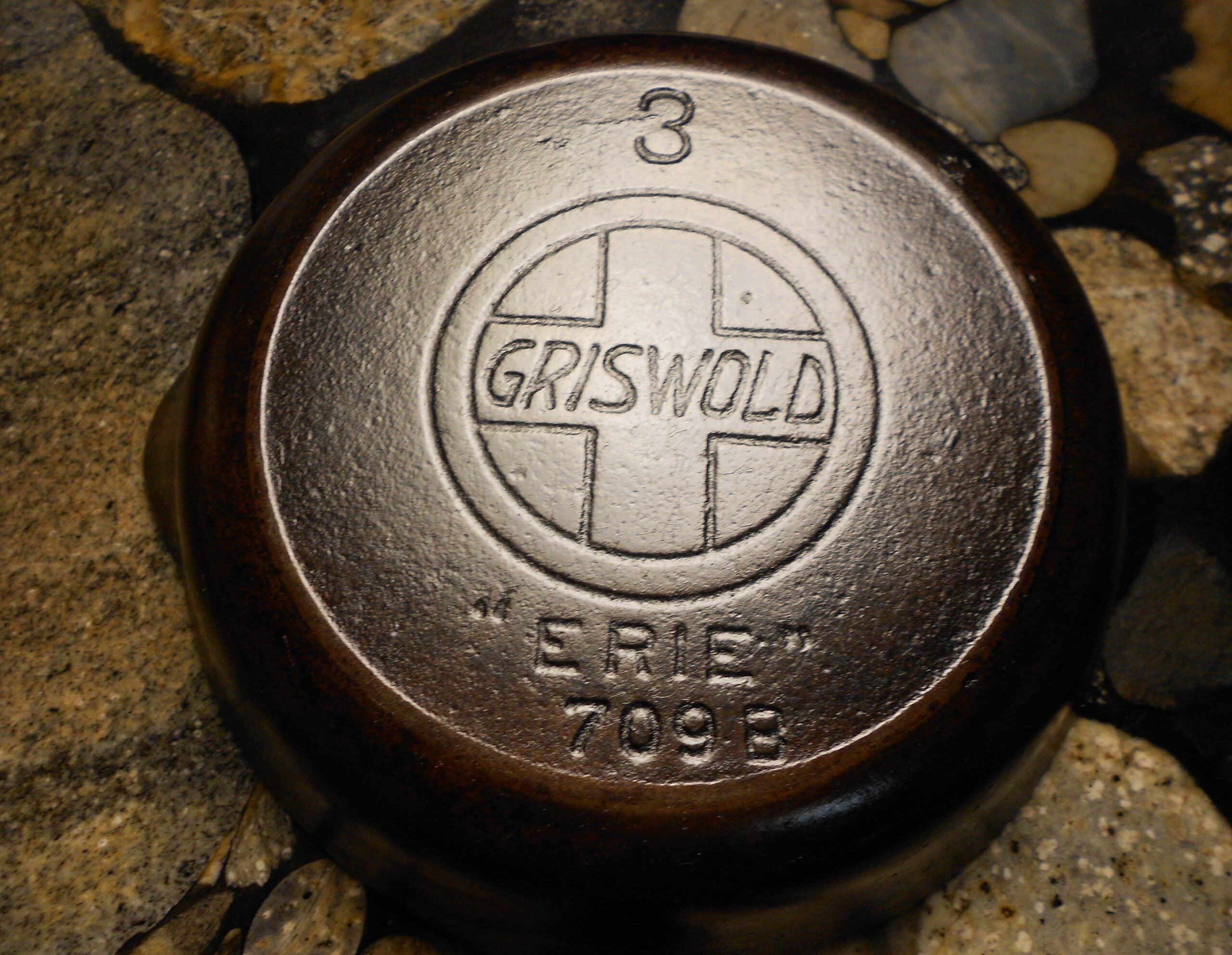 A close-up of the bottom of a vintage cast iron skillet, showing the raised "GRISWOLD" logo, the number 3, "ERIE," and "709 B" on a textured stone surface.