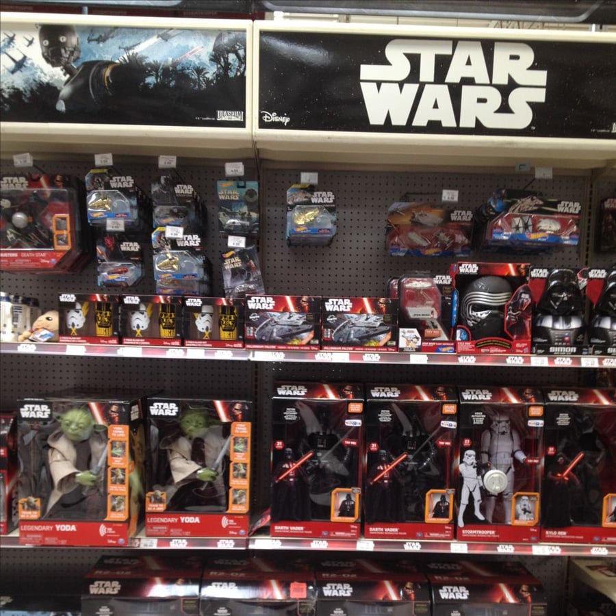 Store shelf filled with various Star Wars action figures, toys, and merchandise, including characters like Yoda, Kylo Ren, and Stormtroopers, with a large Star Wars logo sign above the display.