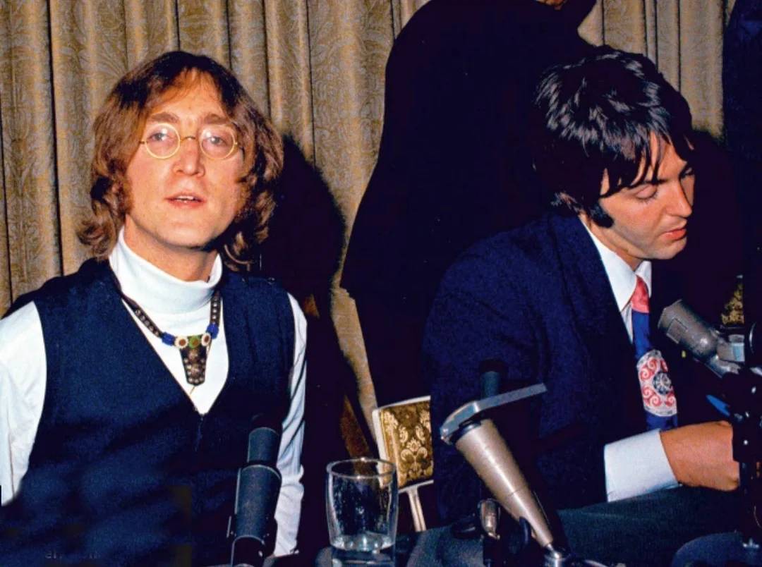 Two men sit at a table with microphones in front of them, appearing to be at a press conference. One wears round glasses, a vest, and a necklace; the other wears a suit and is looking down while writing. A curtain is in the background.