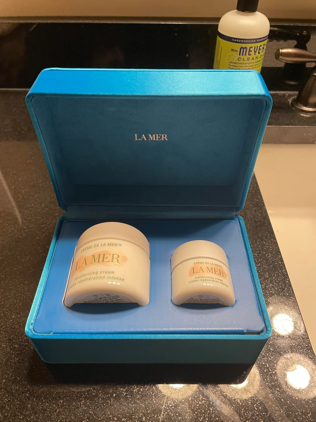 Two jars of La Mer moisturizing cream, one large and one small, are displayed in a blue box with "La Mer" printed inside. The box sits on a black bathroom counter near a bottle of Mrs. Meyer's cleaner and a sink.