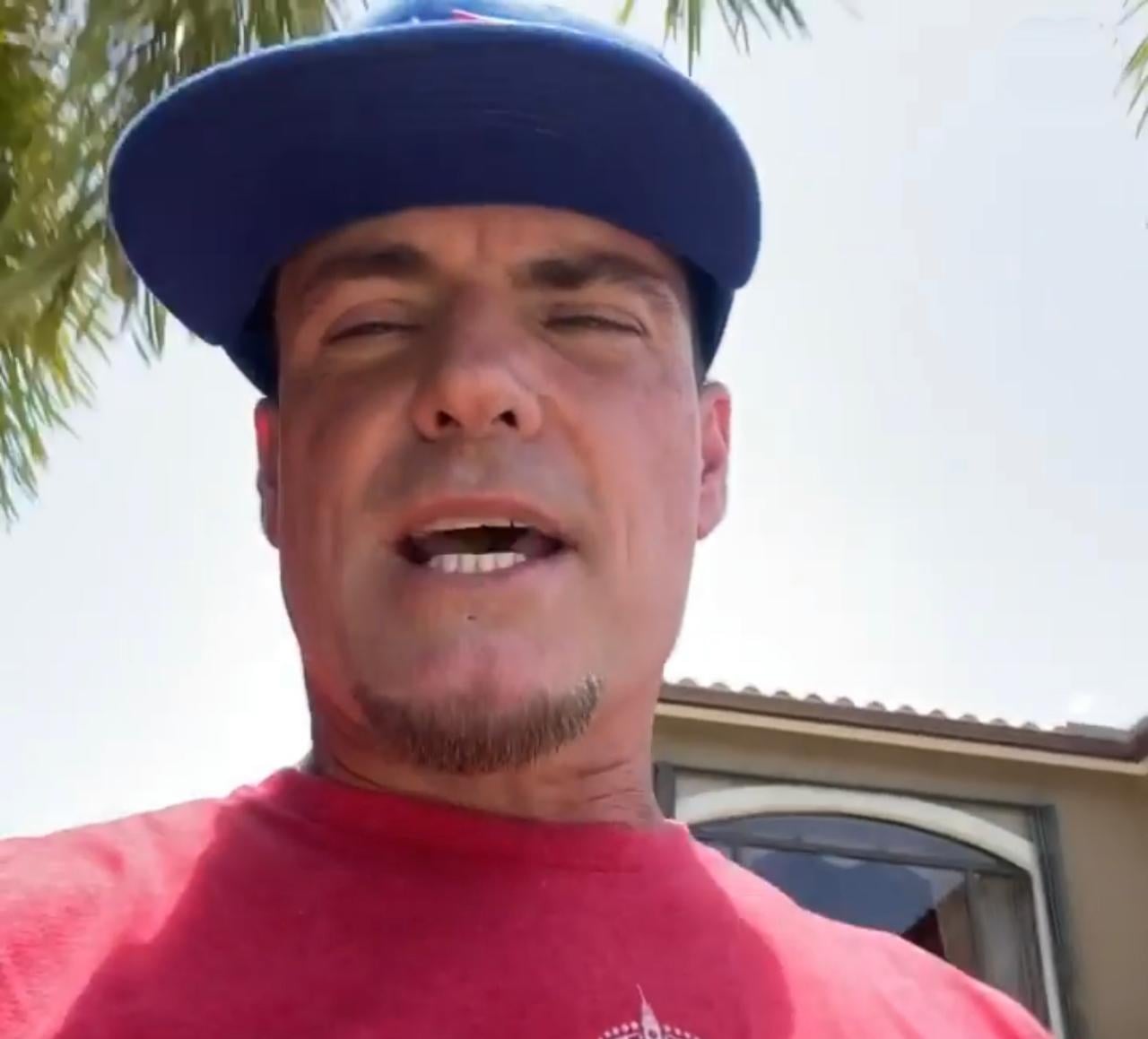 A man wearing a blue baseball cap and red shirt stands outside near a building and palm tree, speaking or singing with his mouth slightly open in bright sunlight.