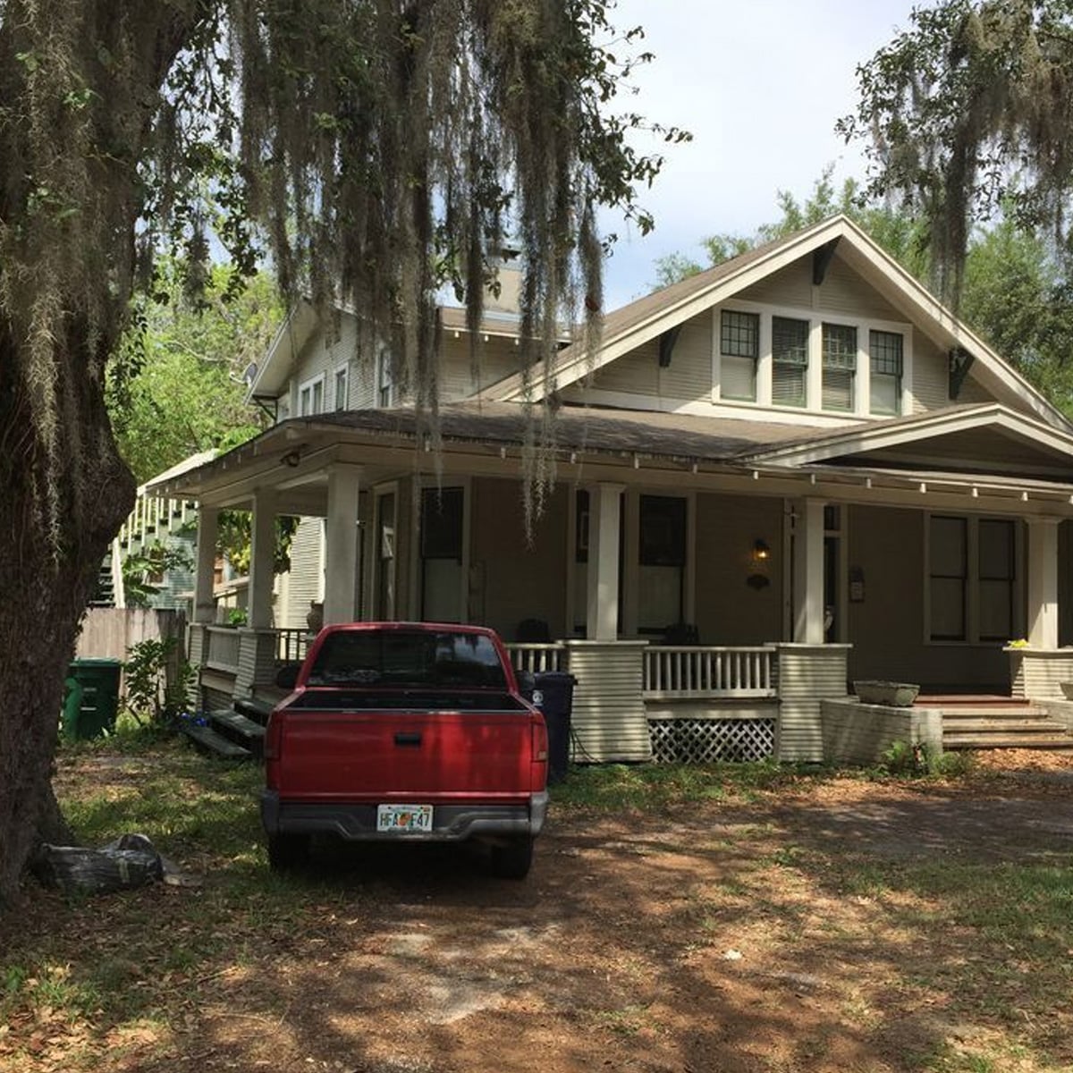 A red pickup truck is parked in front of a large beige house with a wide wraparound porch, surrounded by trees draped with Spanish moss. The yard has patches of grass and dirt.
