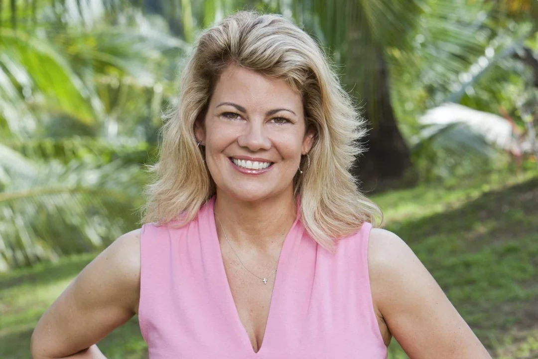A woman with blonde hair wearing a sleeveless pink top stands outdoors, smiling, with green palm trees and grass in the background.