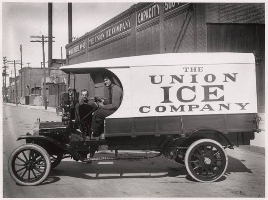 Black-and-white photo of two men sitting in an old-fashioned delivery truck labeled "The Union Ice Company," parked on a street beside a large brick building.