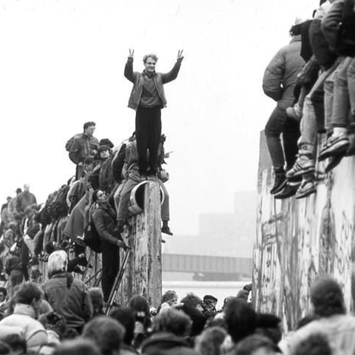 Crowds of people sit and stand on top of the Berlin Wall, some with arms raised in victory, during its fall, symbolizing freedom and unity. The background is hazy with more people and buildings visible.
