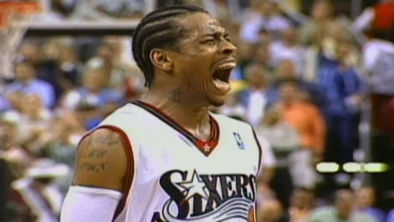 A basketball player in a white Sixers jersey with braided hair and visible tattoos shouts passionately during a game, with a blurred crowd in the background.