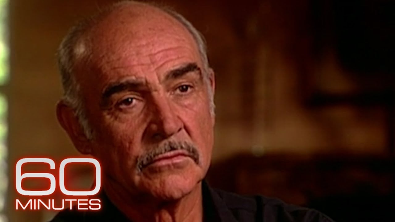 An older man with a mustache and goatee looks serious during an interview. The "60 MINUTES" logo appears in the lower left corner. The background is softly blurred with warm lighting.