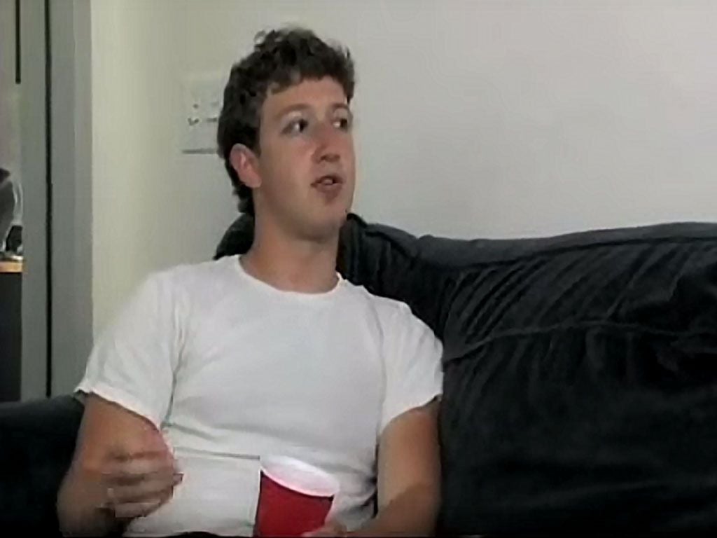 A young man with short curly hair sits on a dark sofa, wearing a plain white t-shirt and holding a red plastic cup, looking slightly to his left in a casual indoor setting.