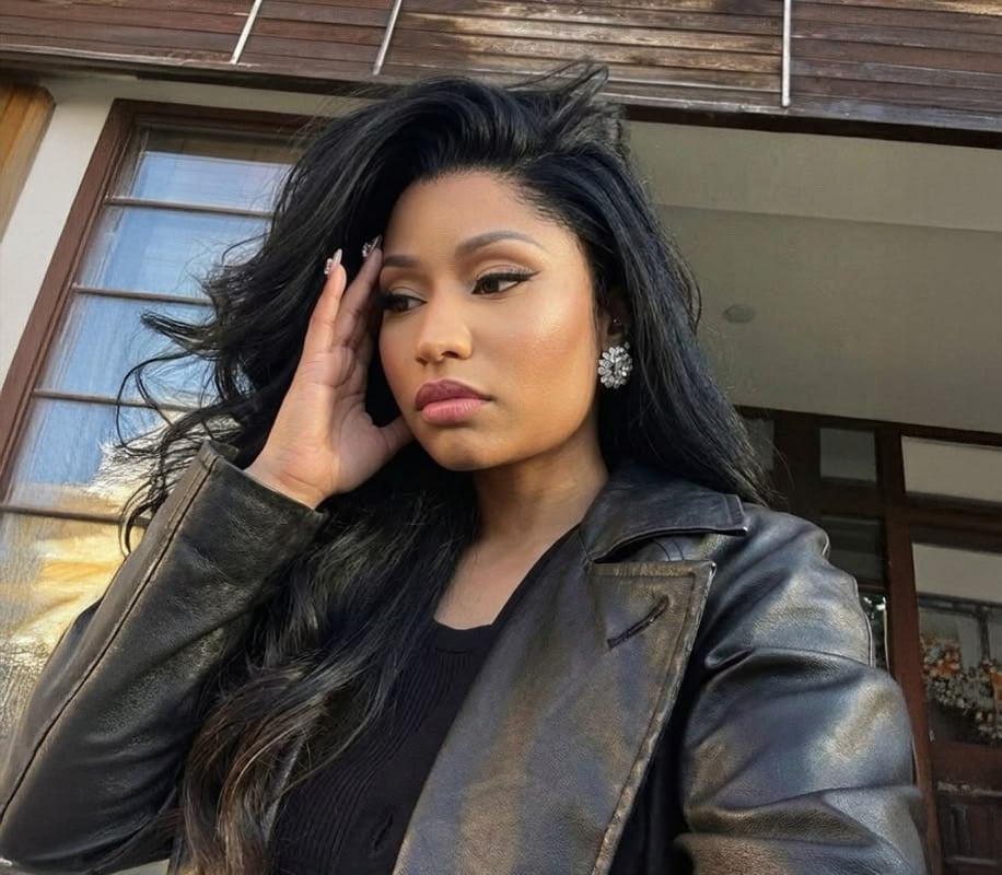 A woman with long black hair wearing a black leather jacket and statement earrings touches her temple while looking thoughtfully to the side, standing in front of a building with large windows.