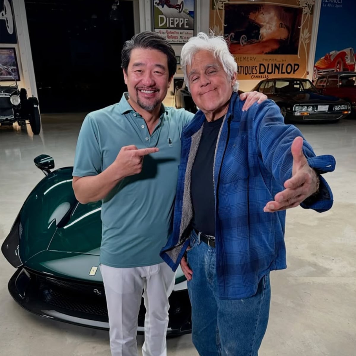 Two smiling men stand in front of a dark green sports car inside a garage with vintage posters on the wall. One man points at the other, who extends his arm towards the camera in a welcoming gesture.