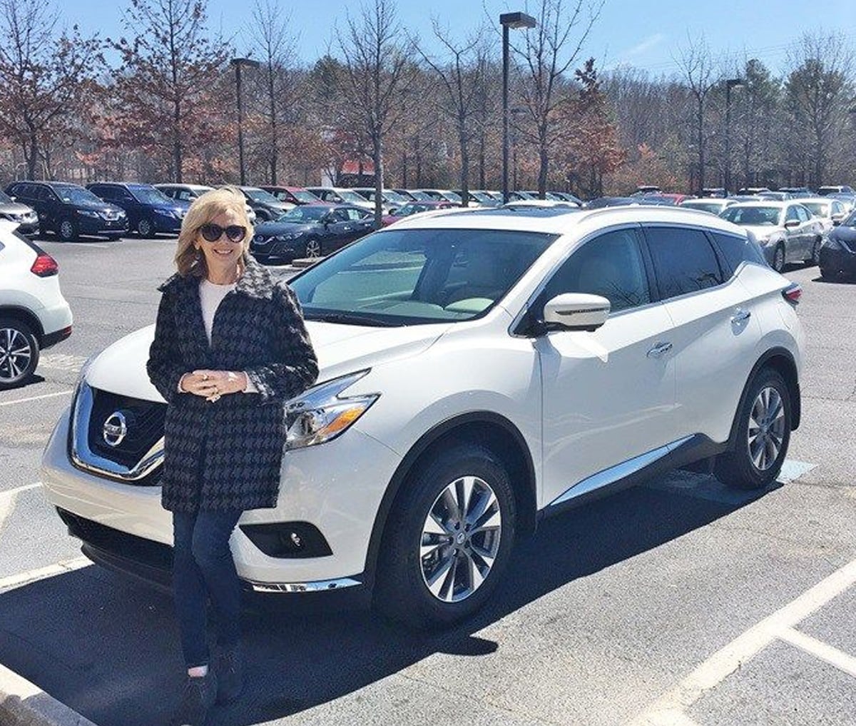 A woman wearing sunglasses and a dark coat stands and smiles in front of a white Nissan SUV parked in a car lot on a sunny day. Other cars and trees are visible in the background.