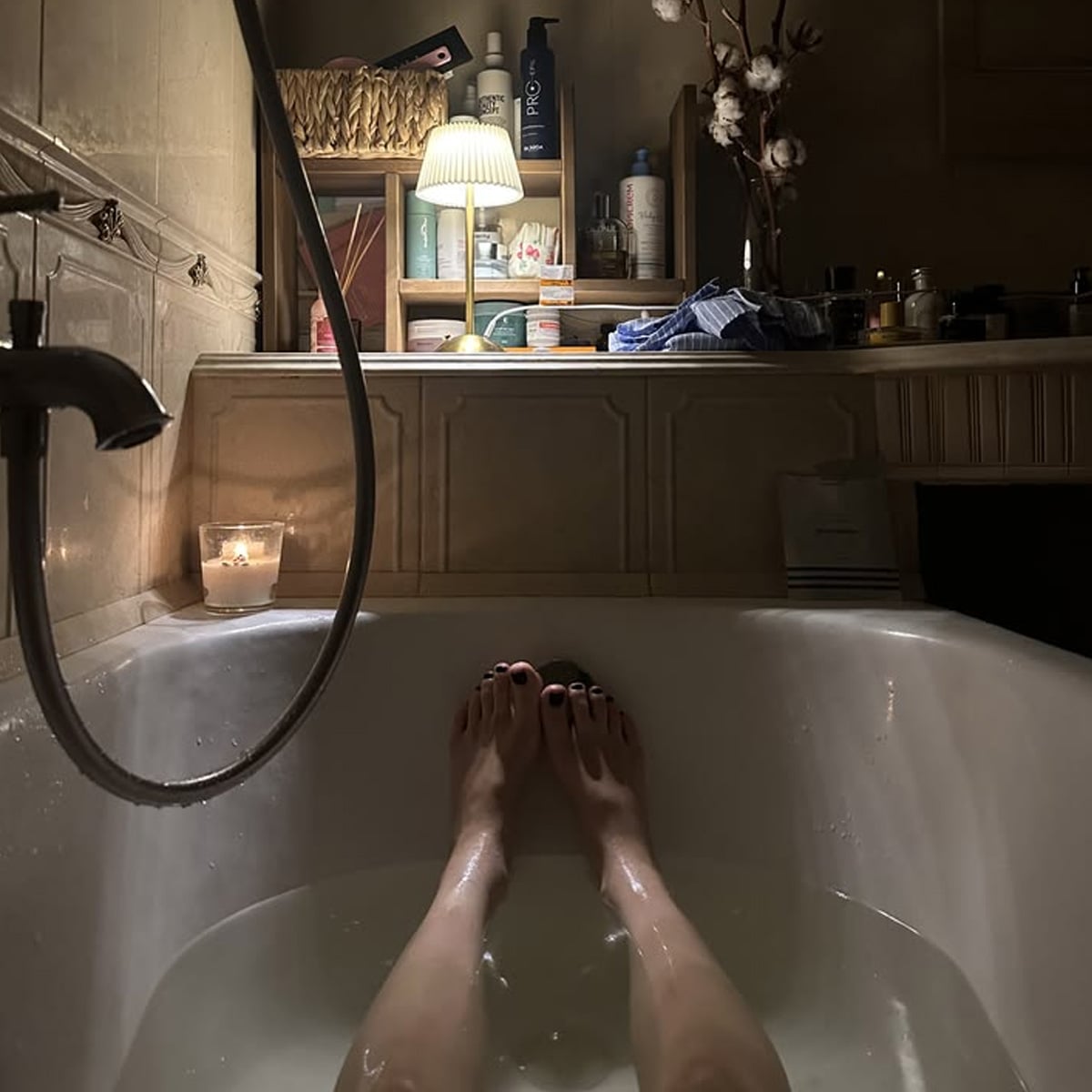 A person relaxes in a dimly lit bathtub with a lit candle nearby. Shelves above the tub hold toiletries, skincare products, and a lamp, creating a cozy and calming atmosphere.