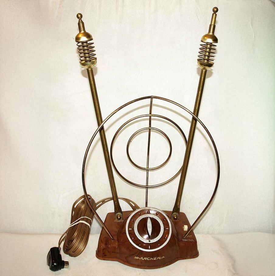 Vintage television antenna with two brass rods, circular metal loops, a central tuning dial, and a brown base. An attached cord with a plug is visible on the left side against a plain background.