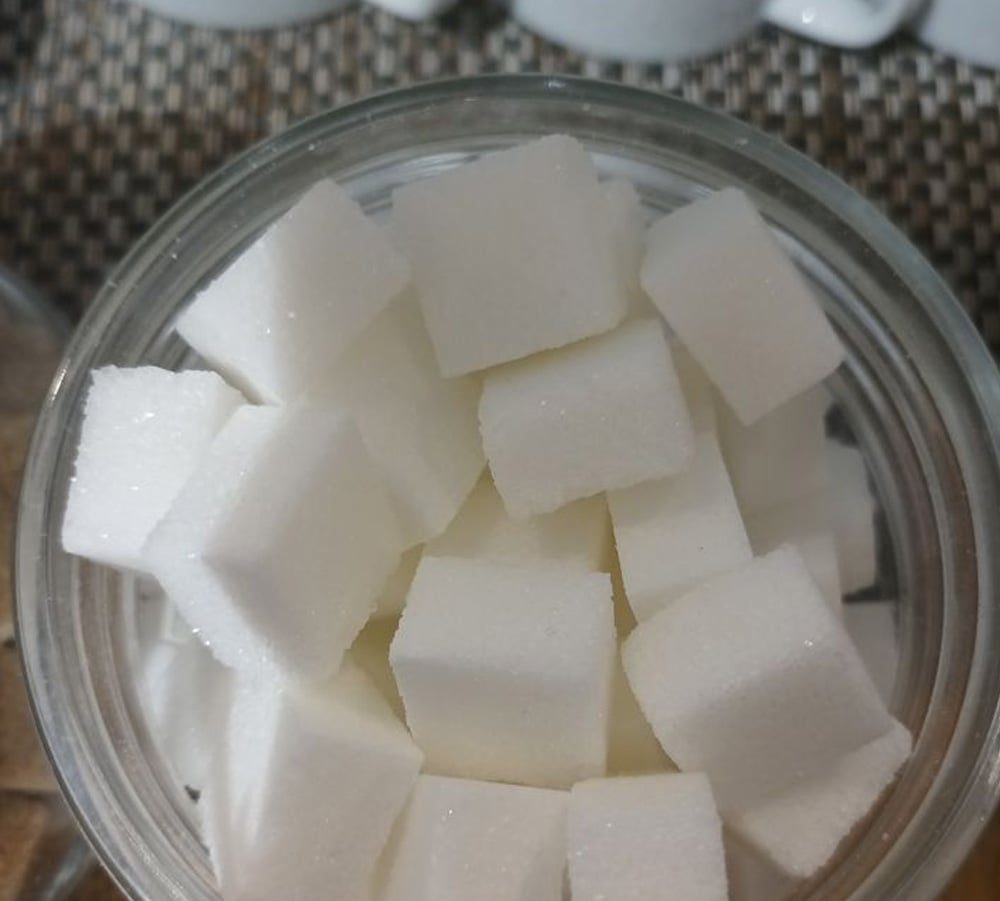 A glass jar filled with white sugar cubes, viewed from above, with part of a woven surface and white objects visible in the background.