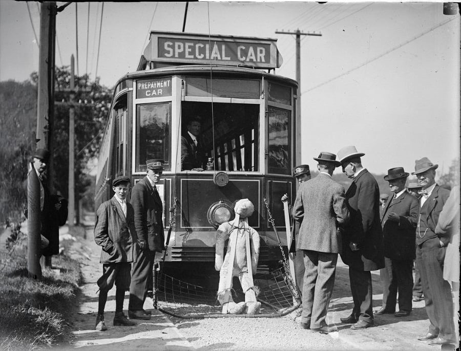 A group of men in early 20th-century attire stand around a streetcar labeled "Special Car." A mannequin or dummy is attached to a safety device on the front of the streetcar, possibly for a demonstration.