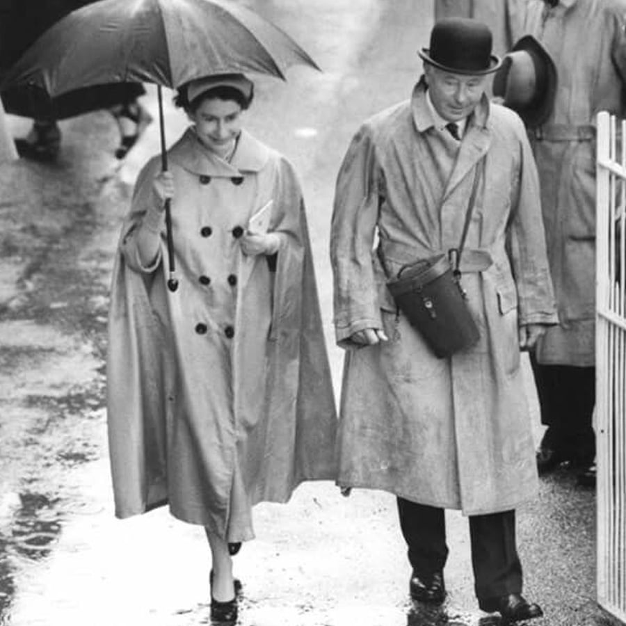 A woman and a man walk side by side in the rain. The woman holds an umbrella over both of them, and they both wear long coats. The man also wears a bowler hat and carries a shoulder bag. The ground appears wet.