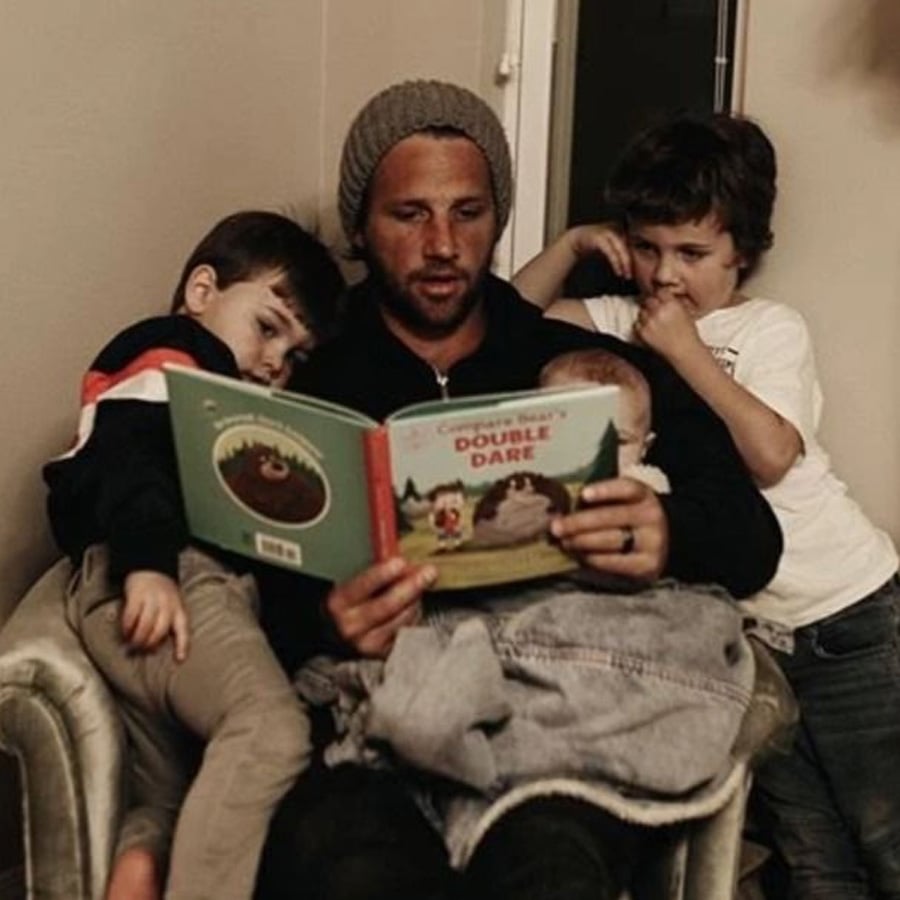 A man wearing a beanie sits in a chair reading a picture book to three young children; two boys are leaning in close, listening, while the third child sits on his lap.