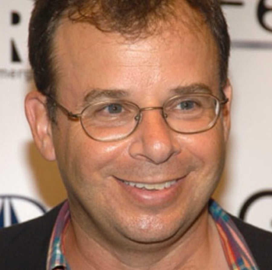 A smiling man with short brown hair, glasses, and a plaid shirt stands in front of a blurred background with text and logos.