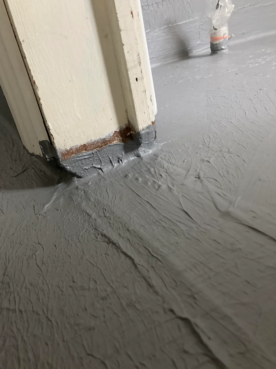 Gray floor paint applied up the base of a white door frame, with some paint covering the bottom of the frame and visible brush or roller marks on the textured surface. A pipe wrapped in plastic is seen in the background.