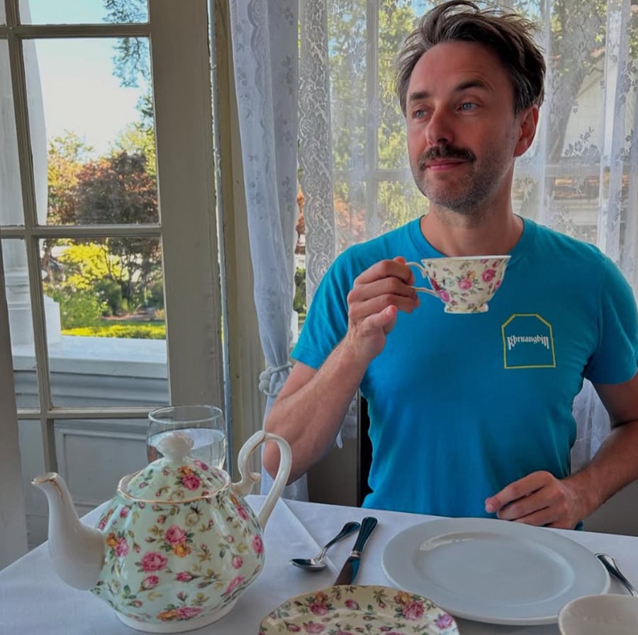 A man with a mustache, wearing a blue T-shirt, sits at a table by a window and holds a floral teacup. There is a matching floral teapot, plate, and glass on the table, with greenery visible outside.