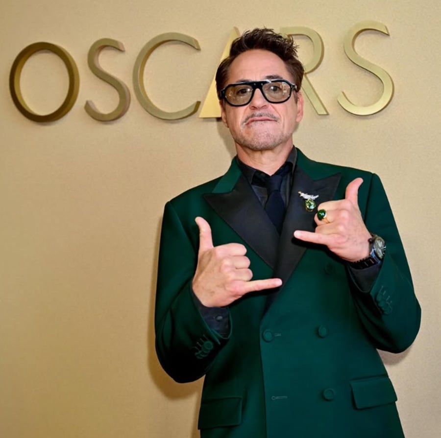 A man in a dark green suit and black shirt poses with shaka hand signs in front of a gold wall with the word "Oscars" partially visible in large letters.