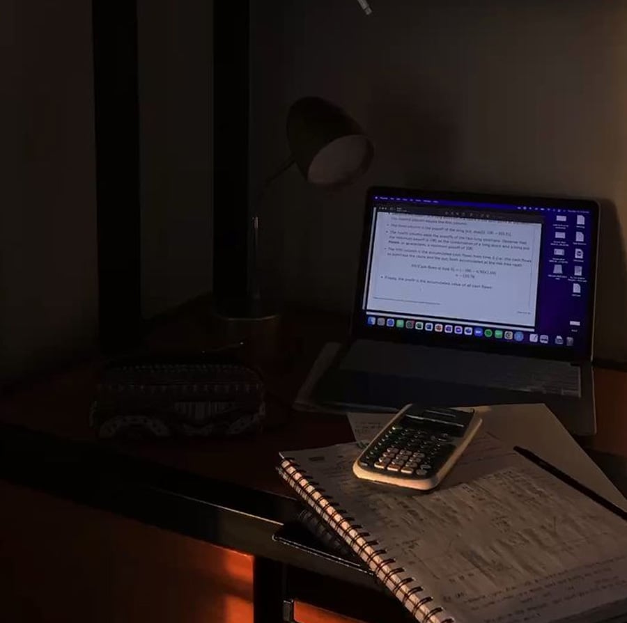 A dimly lit desk with a laptop displaying text, a lamp, a notebook with handwritten notes, a pencil, a calculator, and a pencil case. The scene suggests late-night studying or work.