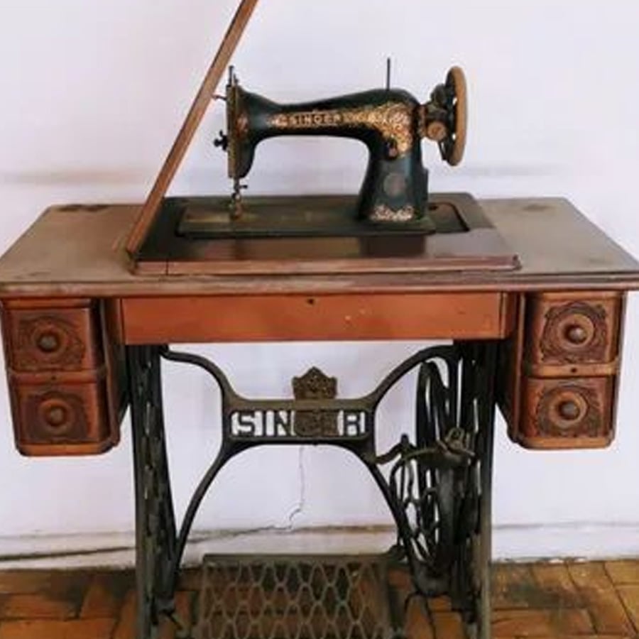 An antique black and gold Singer sewing machine is mounted on a wooden table with four side drawers and a metal treadle base. The machine is partly open, showing its intricate design.