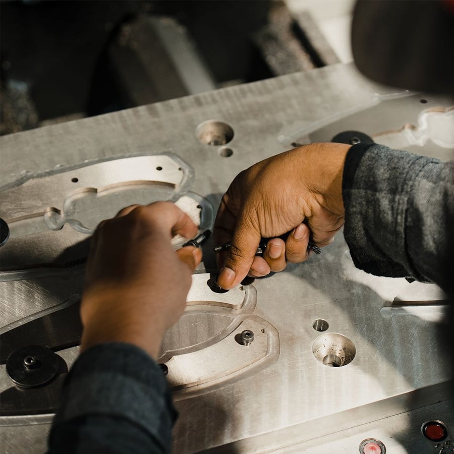 Close-up of a person’s hands using tools to work on a metal machine part with cut-out shapes and screws, likely as part of a manufacturing or assembly process.