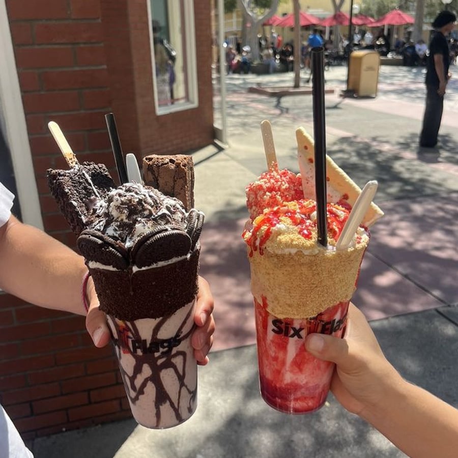 Two people holding elaborate milkshakes outdoors; one is chocolate with Oreos and brownies, the other is strawberry with whipped cream, cake pieces, and sprinkles. Both shakes are topped with various sweets and served in clear cups.