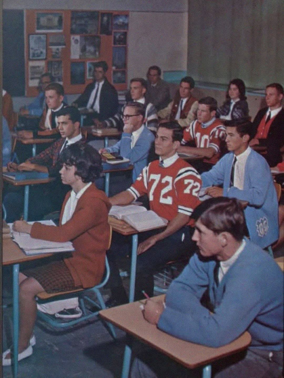A classroom of high school students from the 1960s, some in red varsity sweaters and sports jerseys, sit at desks with notebooks, attentively listening to a lesson. The room has posters and a bulletin board on the back wall.