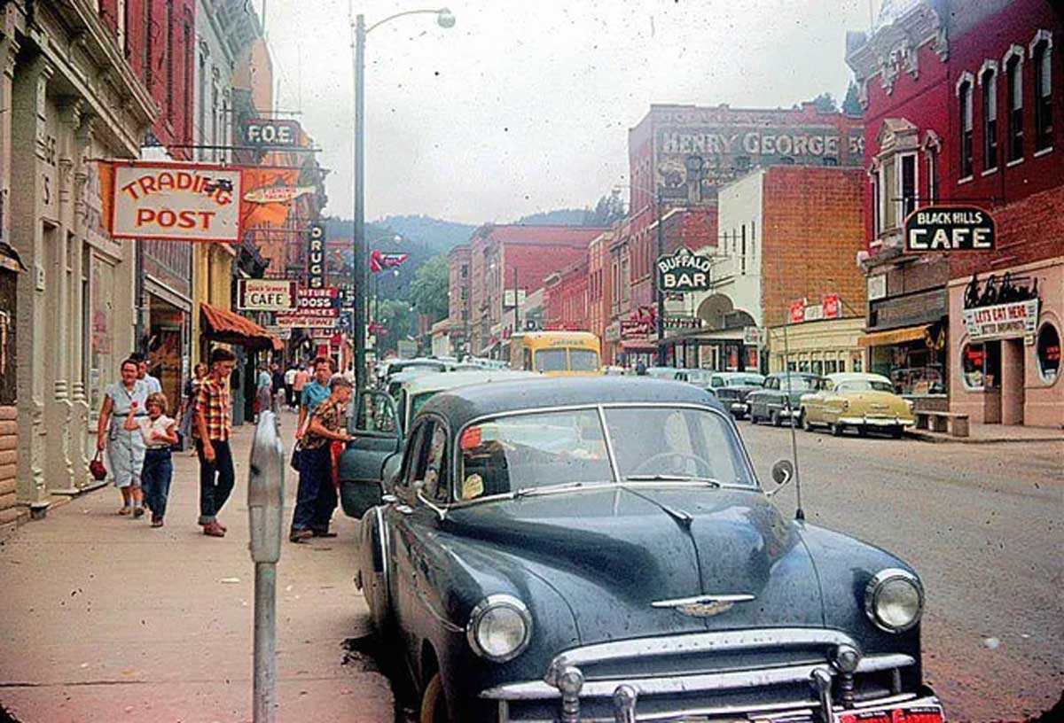 A vintage street scene with classic cars parked along the curb, people walking on the sidewalk, and storefronts with signs for cafes, bars, and shops in a small town.