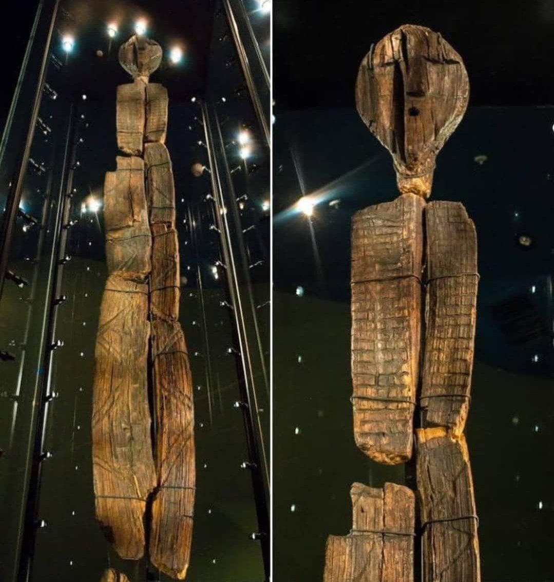 Tall, ancient wooden statue with a carved face and elongated body, displayed upright in a glass case. The surface is cracked and weathered, with faint geometric carvings visible. Two views: full-length and close-up.