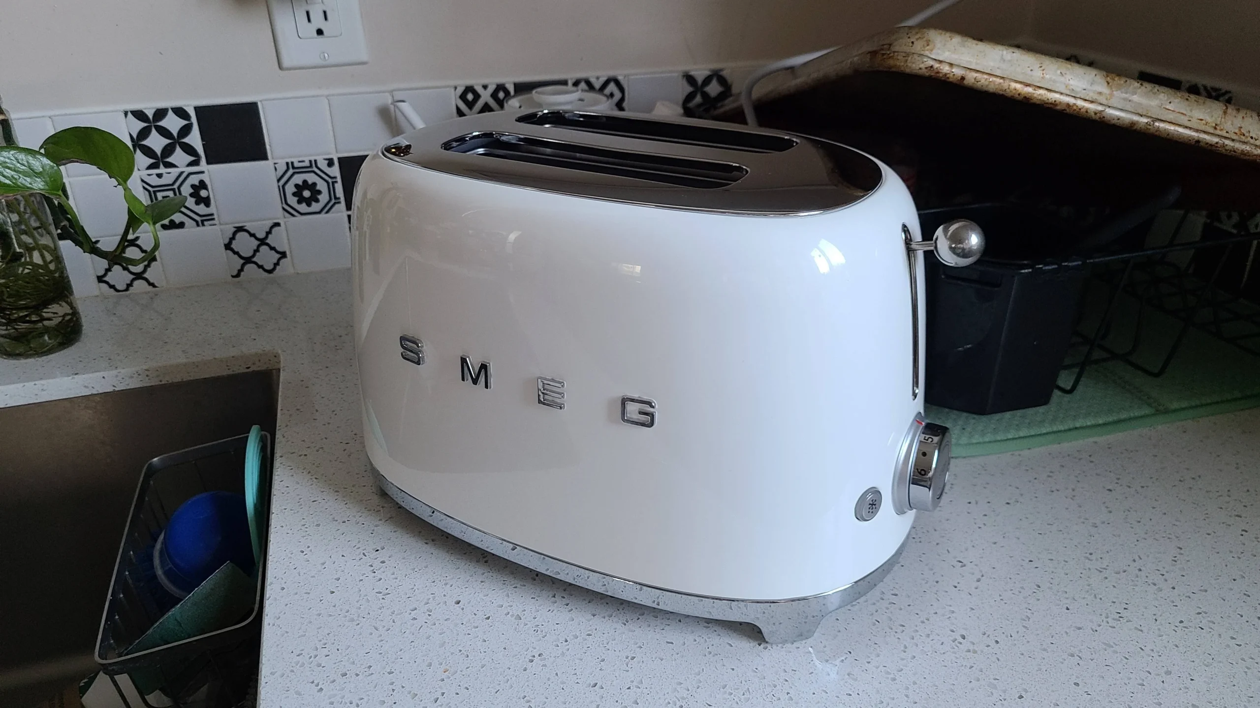 A white SMEG toaster sits on a kitchen countertop near a sink, with patterned black-and-white tiles on the wall and a tray resting nearby.