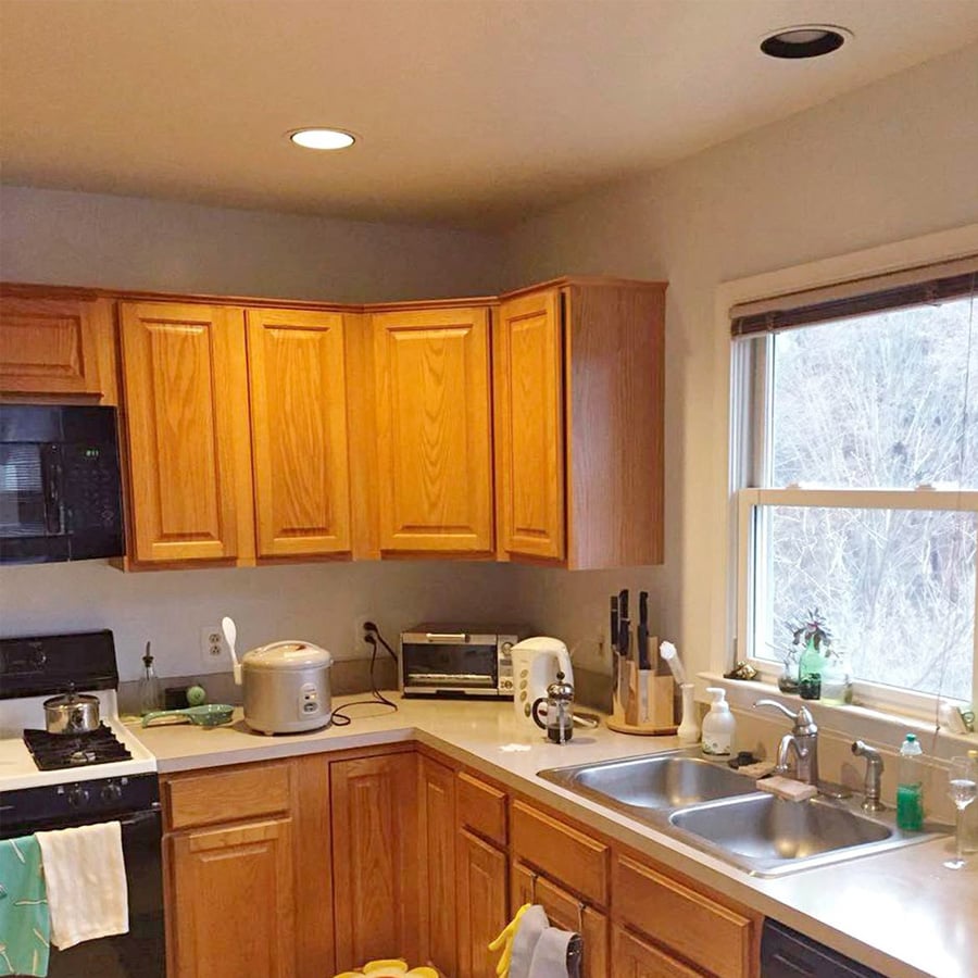 A kitchen with wooden cabinets, a double sink, and a window. Appliances include a microwave, rice cooker, toaster oven, and kettle. Various kitchen items are on the countertops, and trees are visible outside the window.