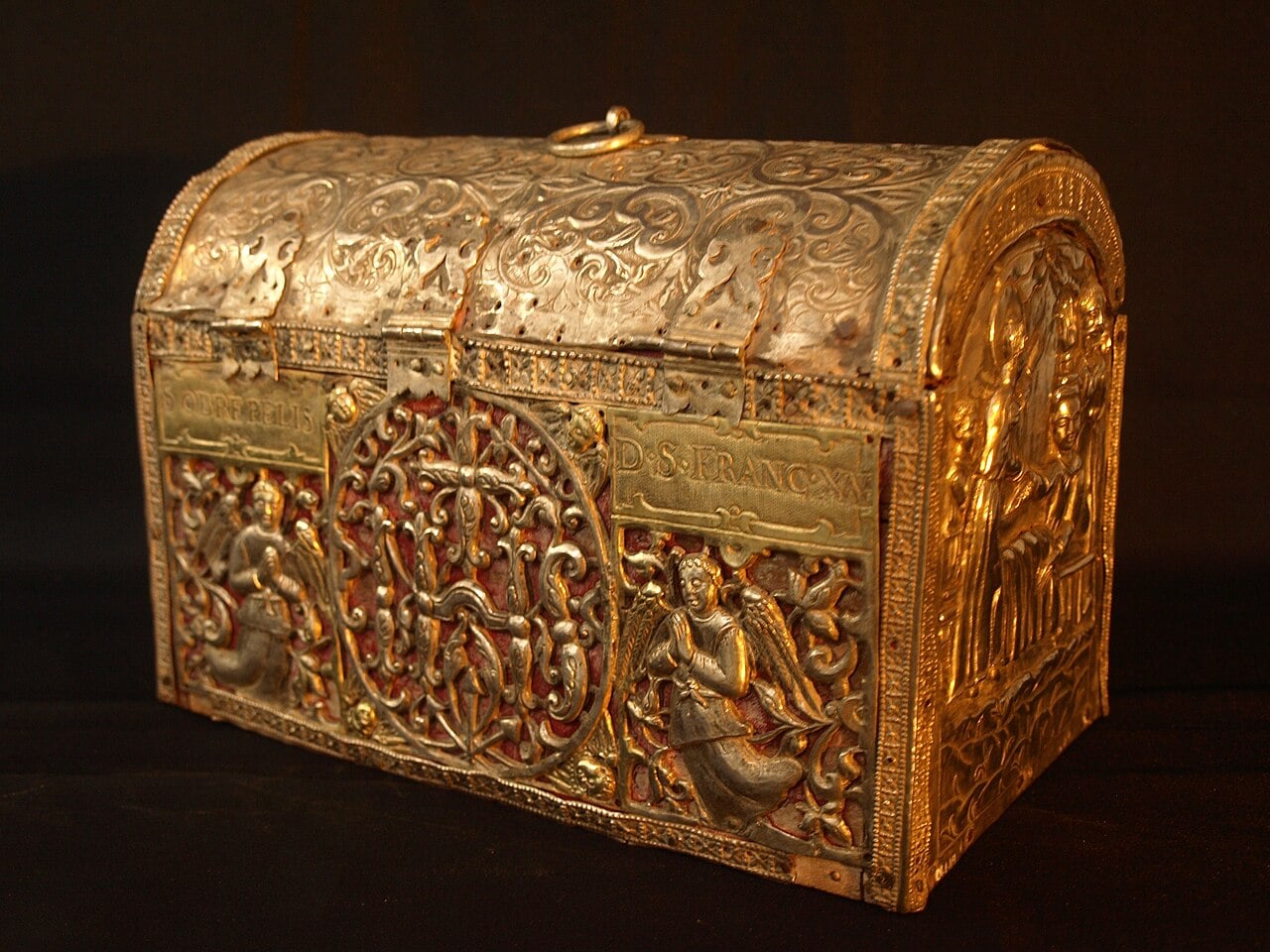 An ornate, antique gold and silver chest with intricate carvings, religious figures, and angels on its sides, set against a black background. The chest has a curved lid and decorative patterns throughout.
