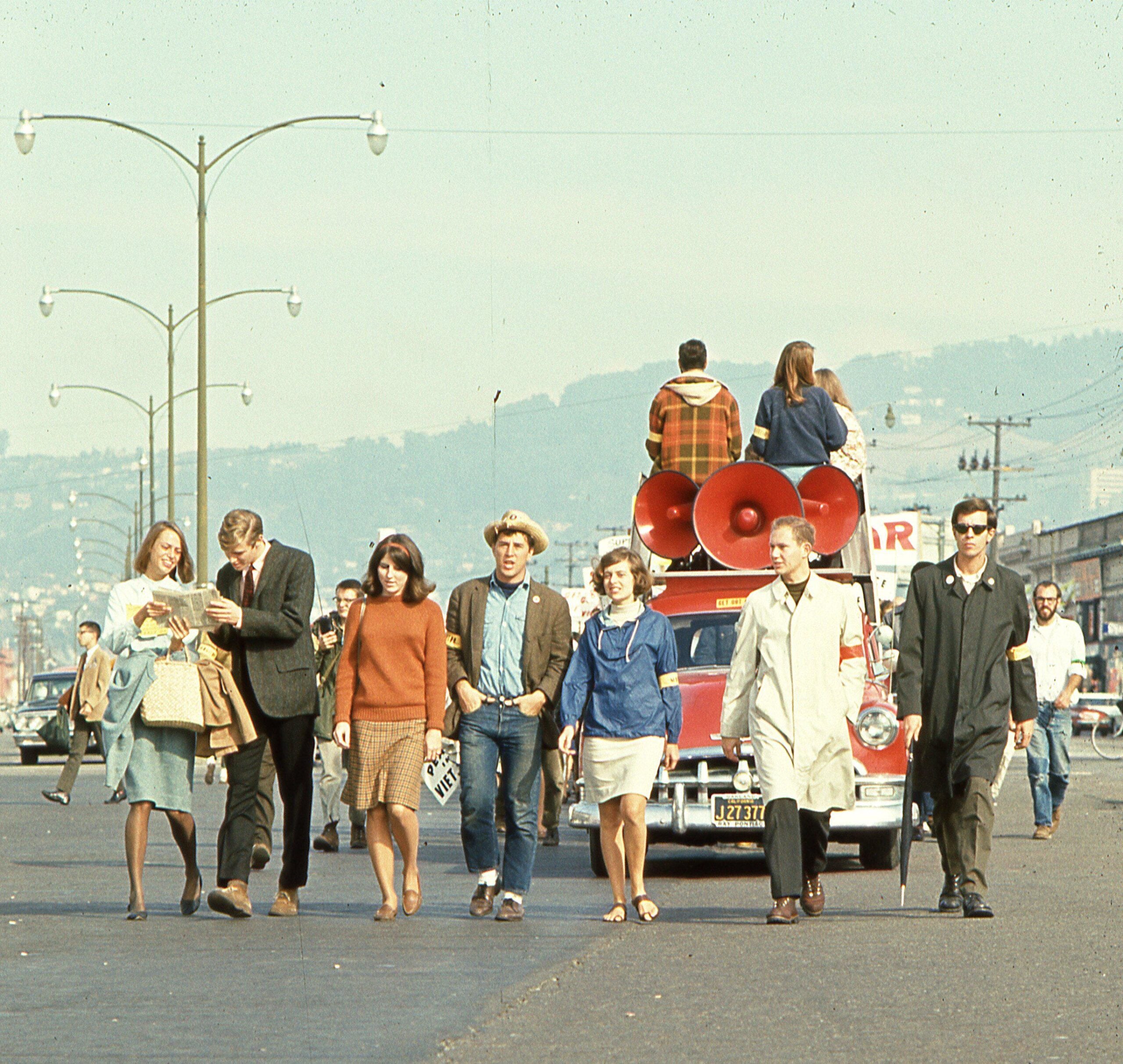 A group of people walk together down a city street, with a vehicle featuring large red loudspeakers parked behind them. Some women wear dresses and coats; a man wears a cowboy hat. Other people sit on top of the vehicle.