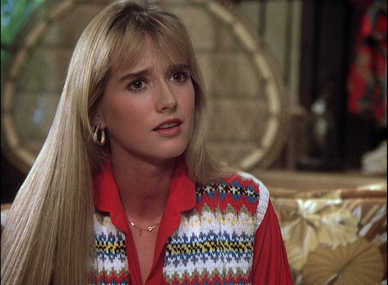 A woman with long blonde hair wearing a colorful patterned sweater vest over a red shirt looks concerned while sitting indoors.