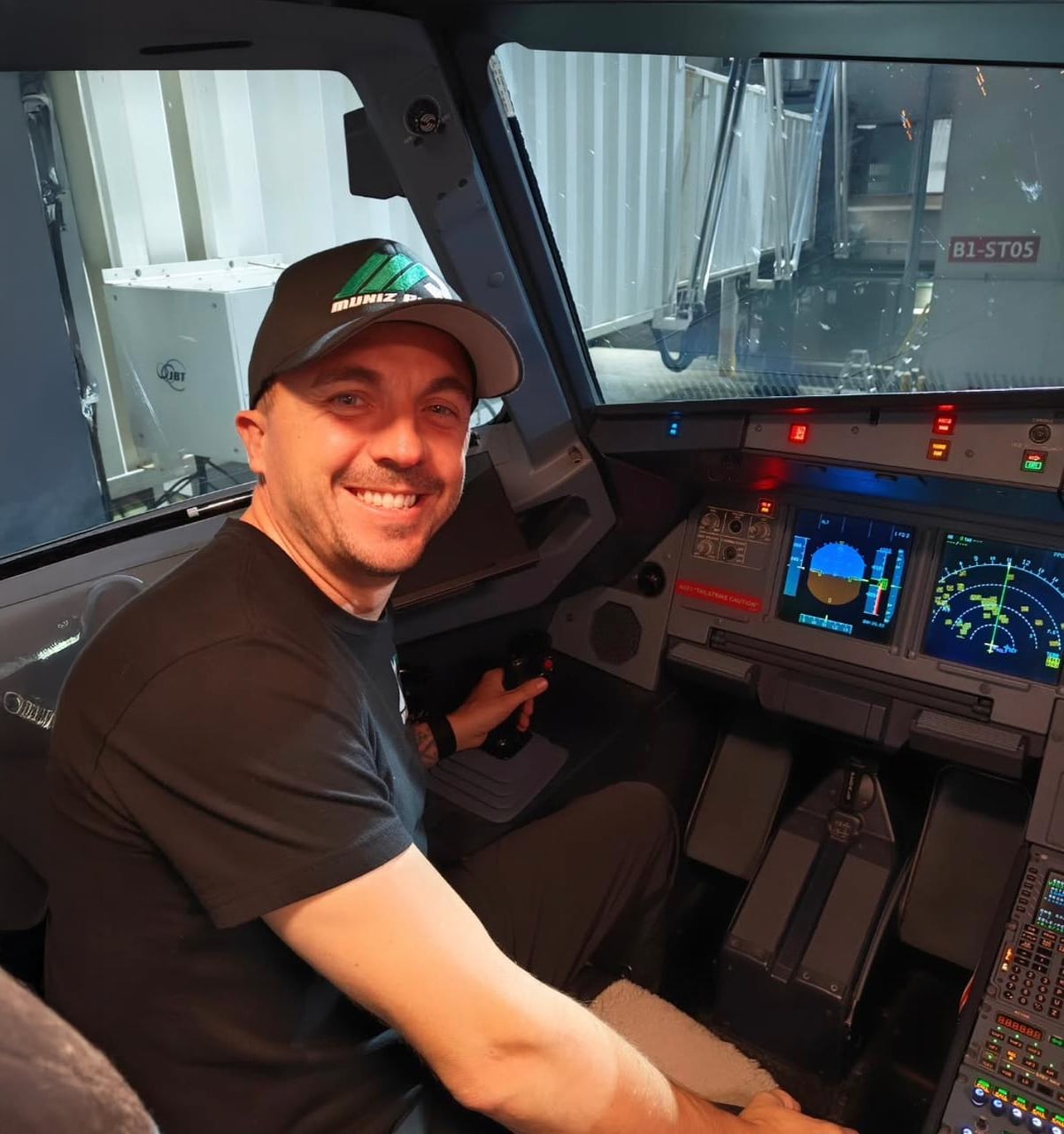 A man wearing a black shirt and hat is smiling while sitting in an airplane cockpit, with various flight instruments and screens visible around him.