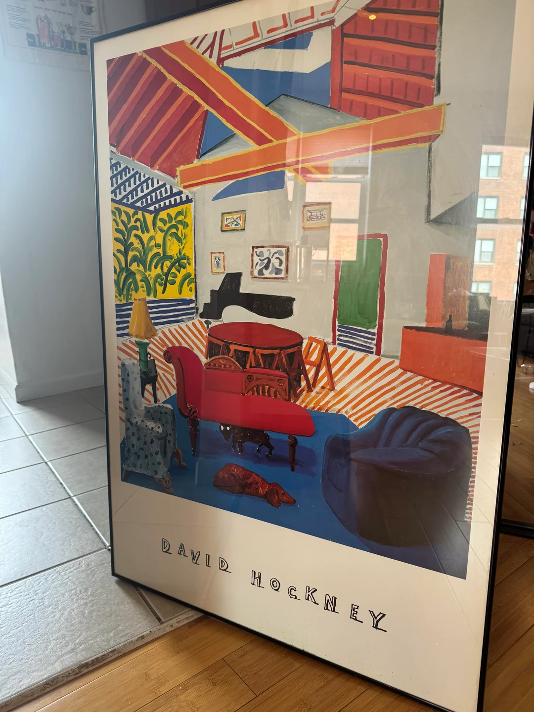 Framed David Hockney art print leaning against a wall, showing a colorful, stylized interior scene with vibrant furniture, patterns, and a piano. The artist’s name is printed at the bottom.