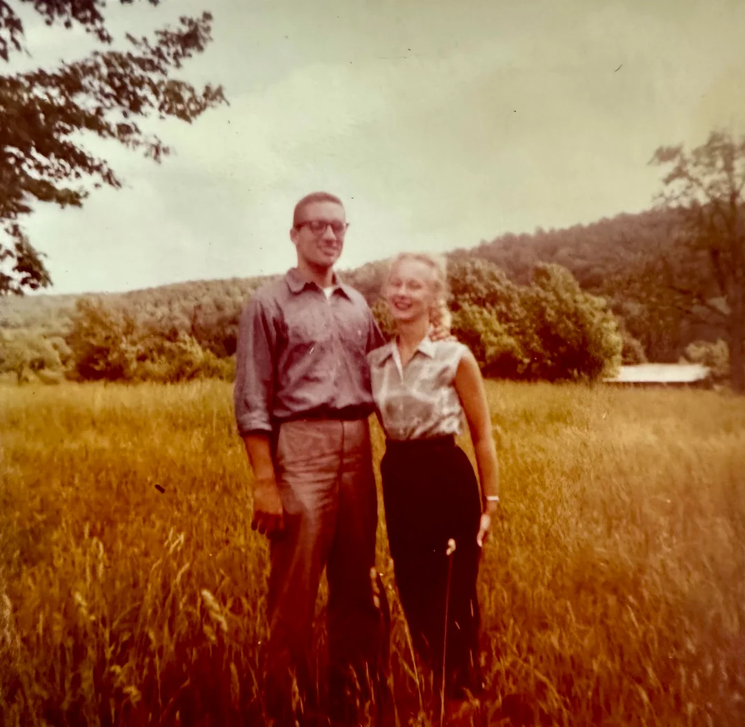 A man and woman stand together in a grassy field with trees and hills in the background. The man wears glasses and a button-up shirt; the woman has light hair and a sleeveless blouse. They both smile at the camera.