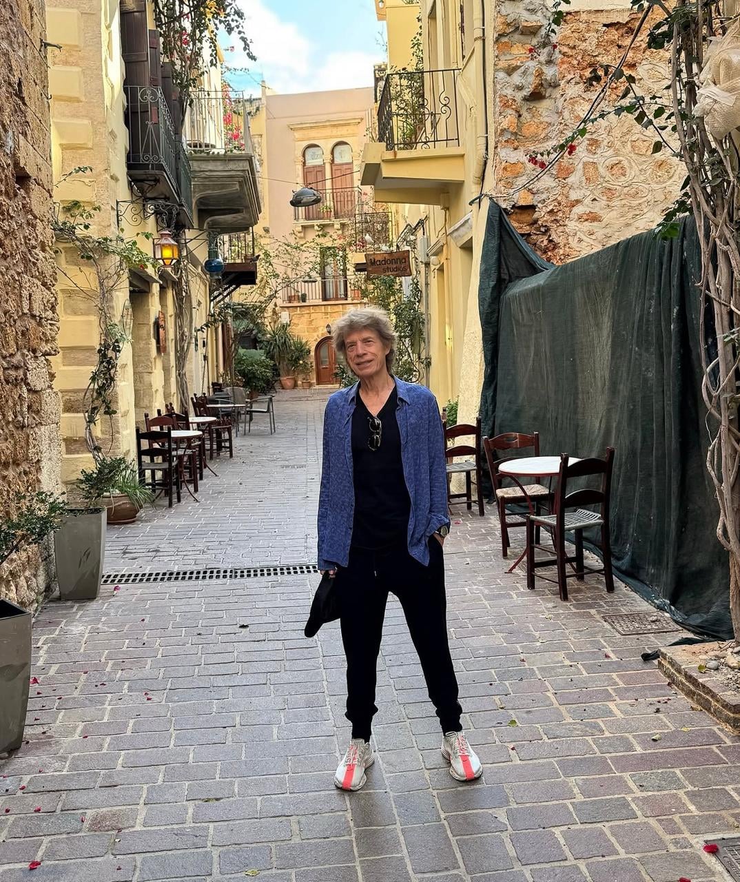 A person with short hair stands in a narrow, cobblestone alley lined with old stone buildings, balconies, and outdoor tables. The person wears a blue jacket, black outfit, and white sneakers. The street is quiet and empty.