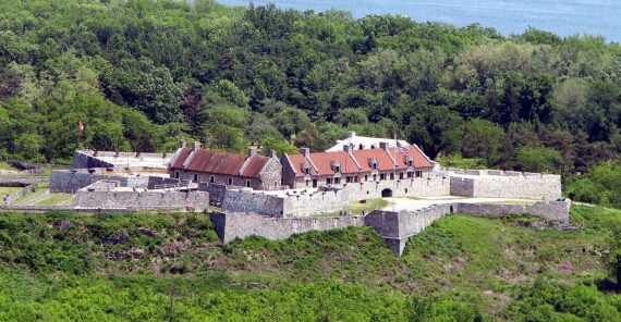 A large stone fort with red-roofed buildings surrounded by green trees sits near the edge of a river, with hills and forested land visible across the water.
