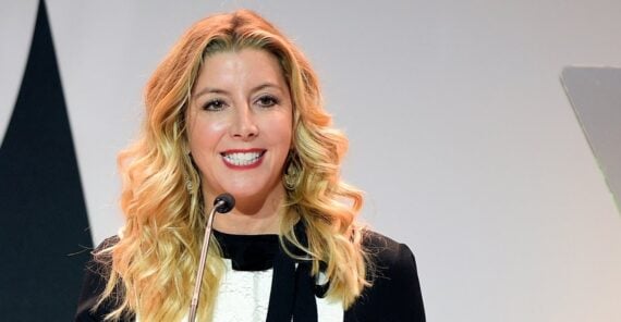 A woman with long, wavy blonde hair smiles while speaking at a podium. She is wearing a black and white outfit and earrings, standing in front of a plain background.