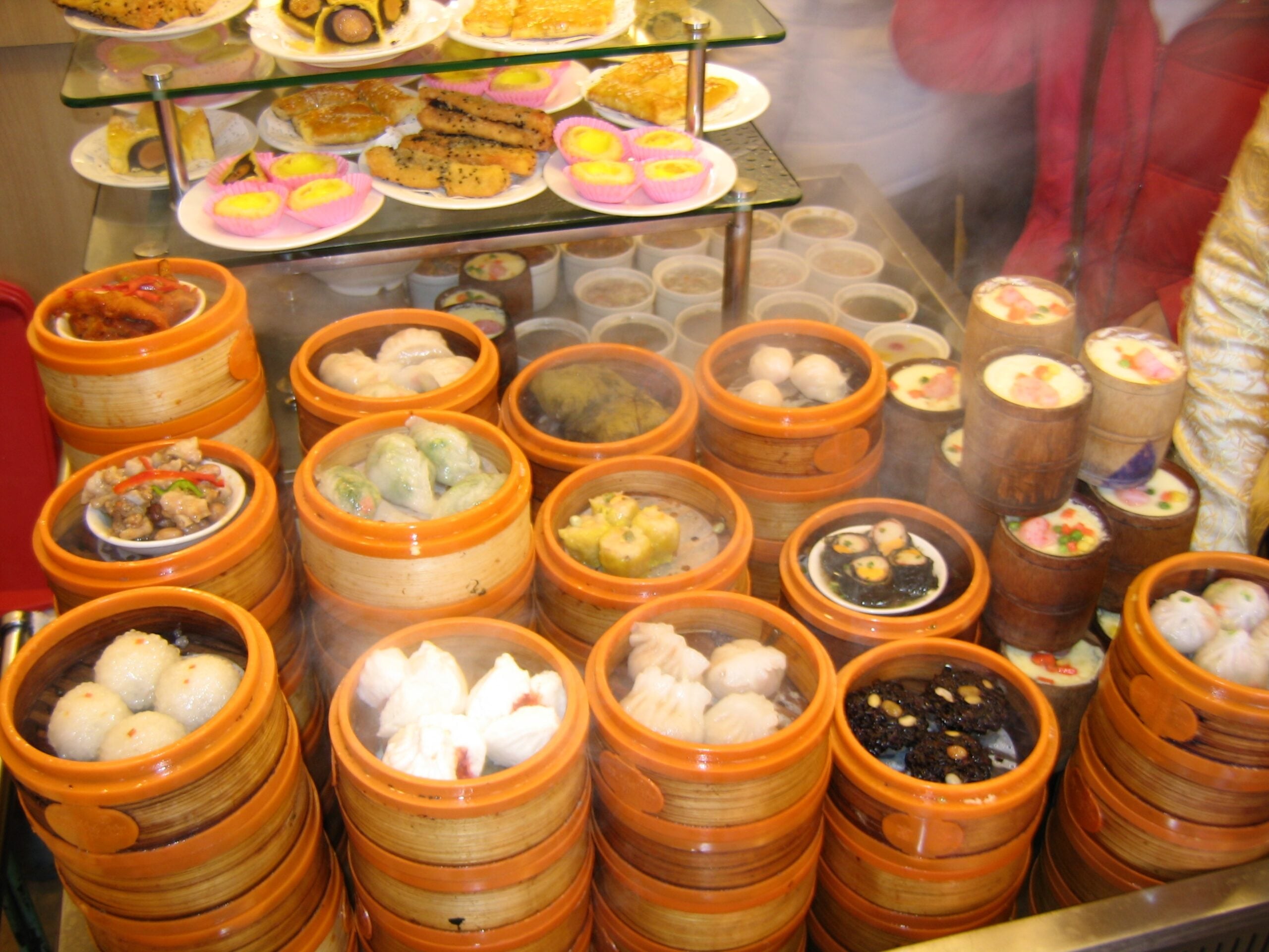 A variety of dim sum dishes in bamboo steamers are displayed on a counter, with steam rising. Colorful pastries and desserts are arranged on glass shelves above the dim sum.