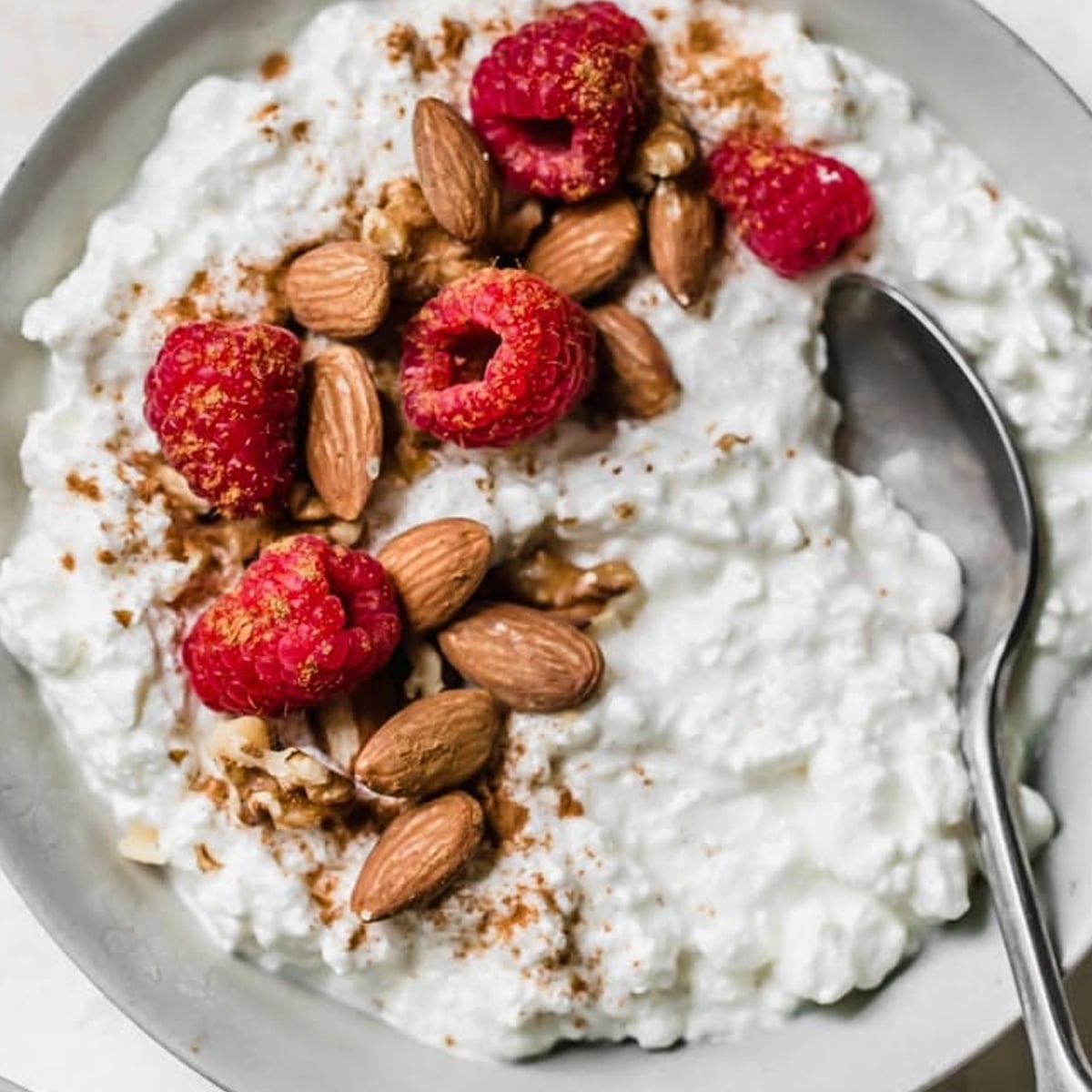 A bowl of cottage cheese topped with whole almonds, raspberries, a sprinkle of cinnamon, and a spoon resting on the side.
