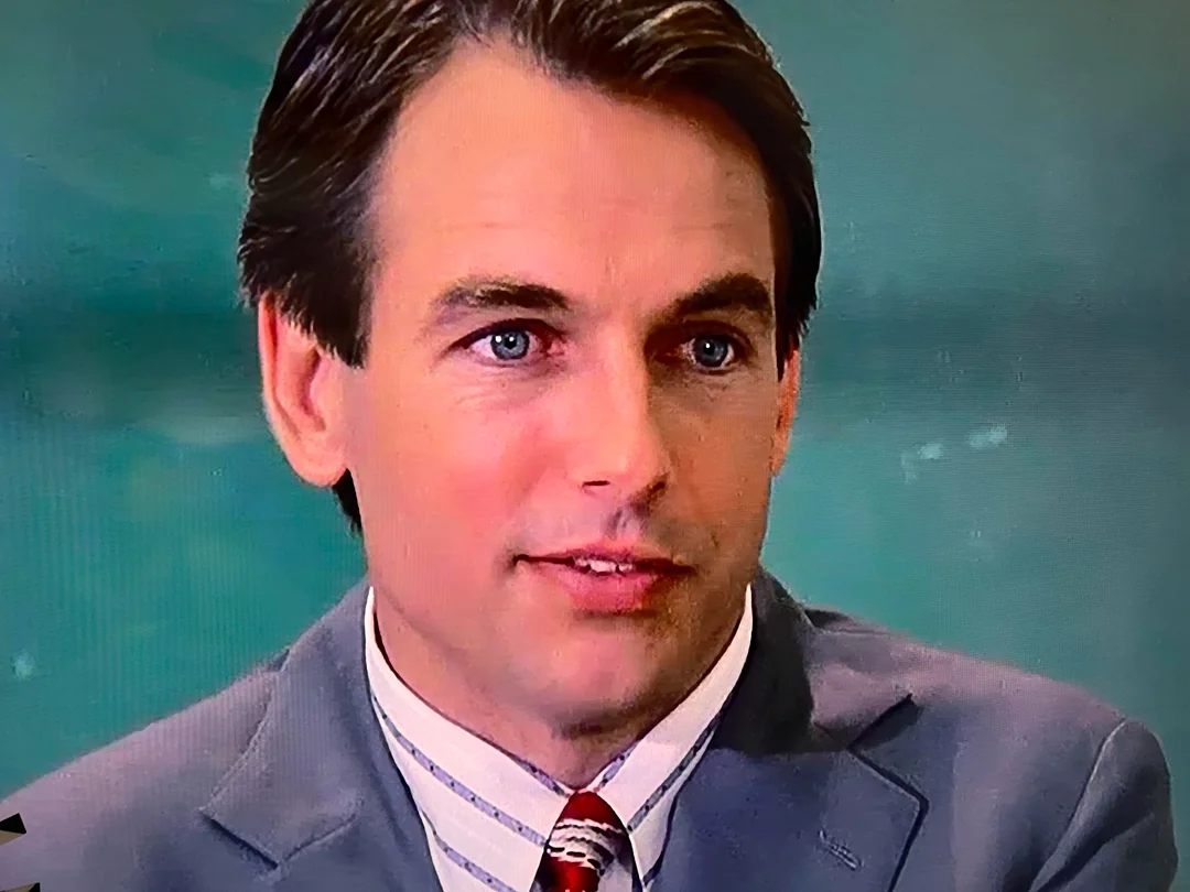 A young man with neatly combed brown hair, wearing a gray suit, white shirt, and striped tie, is looking slightly to the side. The background is blurred in shades of green.
