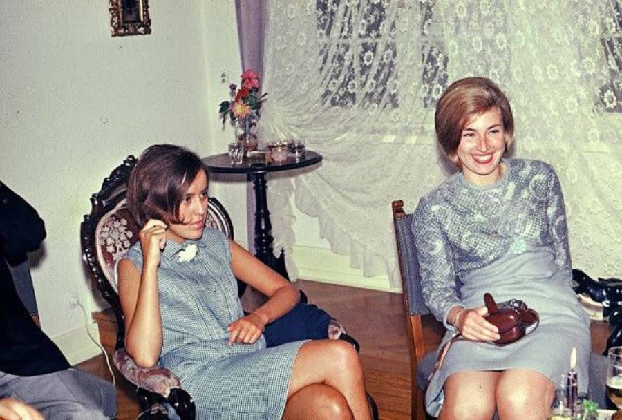 Two women in 1960s-style dresses sit indoors on ornate chairs. One woman is holding a cigarette, and the other is smiling while holding a brown teapot. Lace curtains and a small table with flowers are visible in the background.