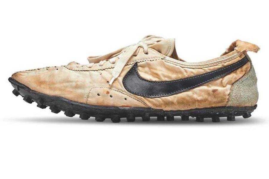 A worn beige Nike running shoe with a black swoosh logo, visible stitching, and a rugged black sole. The shoe appears vintage and distressed, with creases and marks on the fabric.