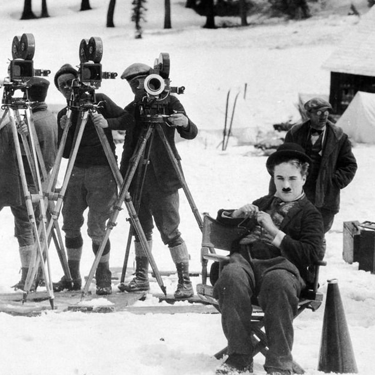 A man in a bowler hat and mustache sits in a director's chair on a snowy film set while three cameramen stand behind vintage film cameras on tripods. Other crew members and equipment are visible.